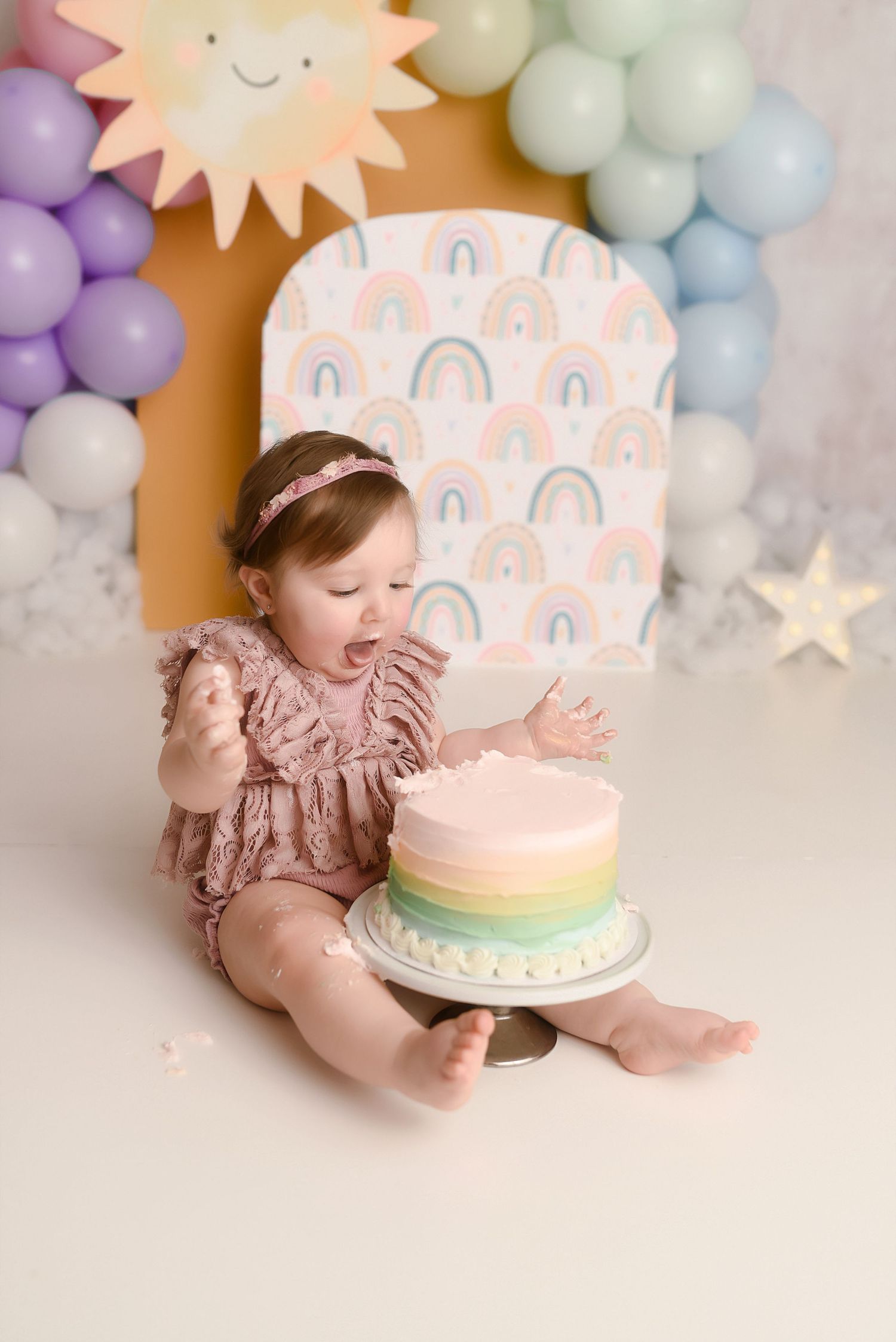 Baby girl in pink romper excitedly smashing her first birthday rainbow cake surrounded by colorful balloons.