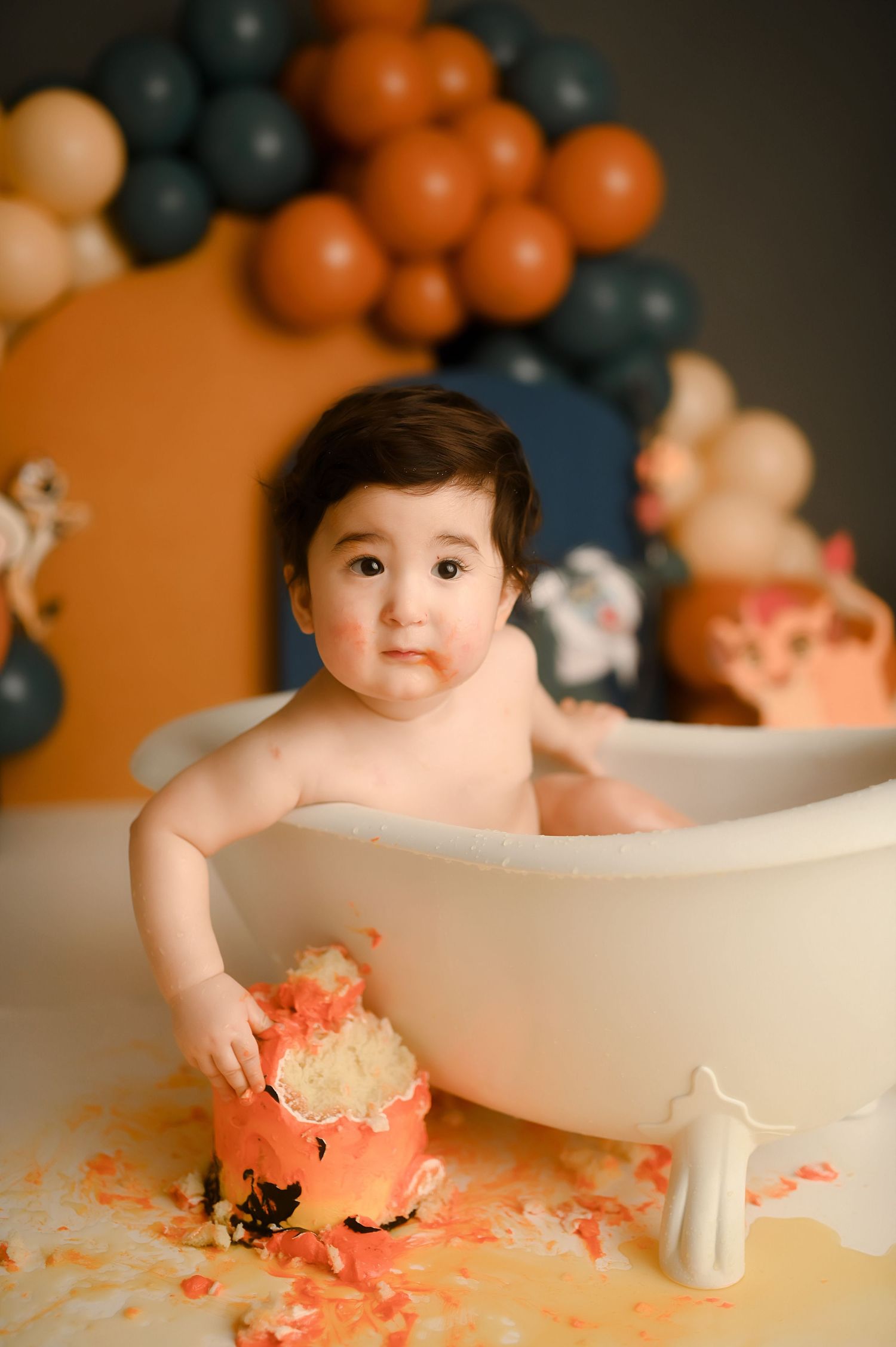 Baby in white tub smashing orange cake, surrounded by teal and orange balloon garland backdrop.
