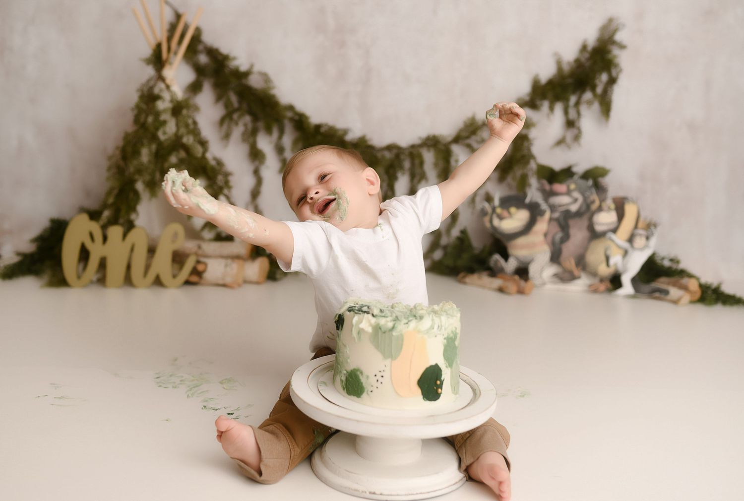 Joyful baby boy raises arms beside green leaf cake at woodland first birthday cake smash.