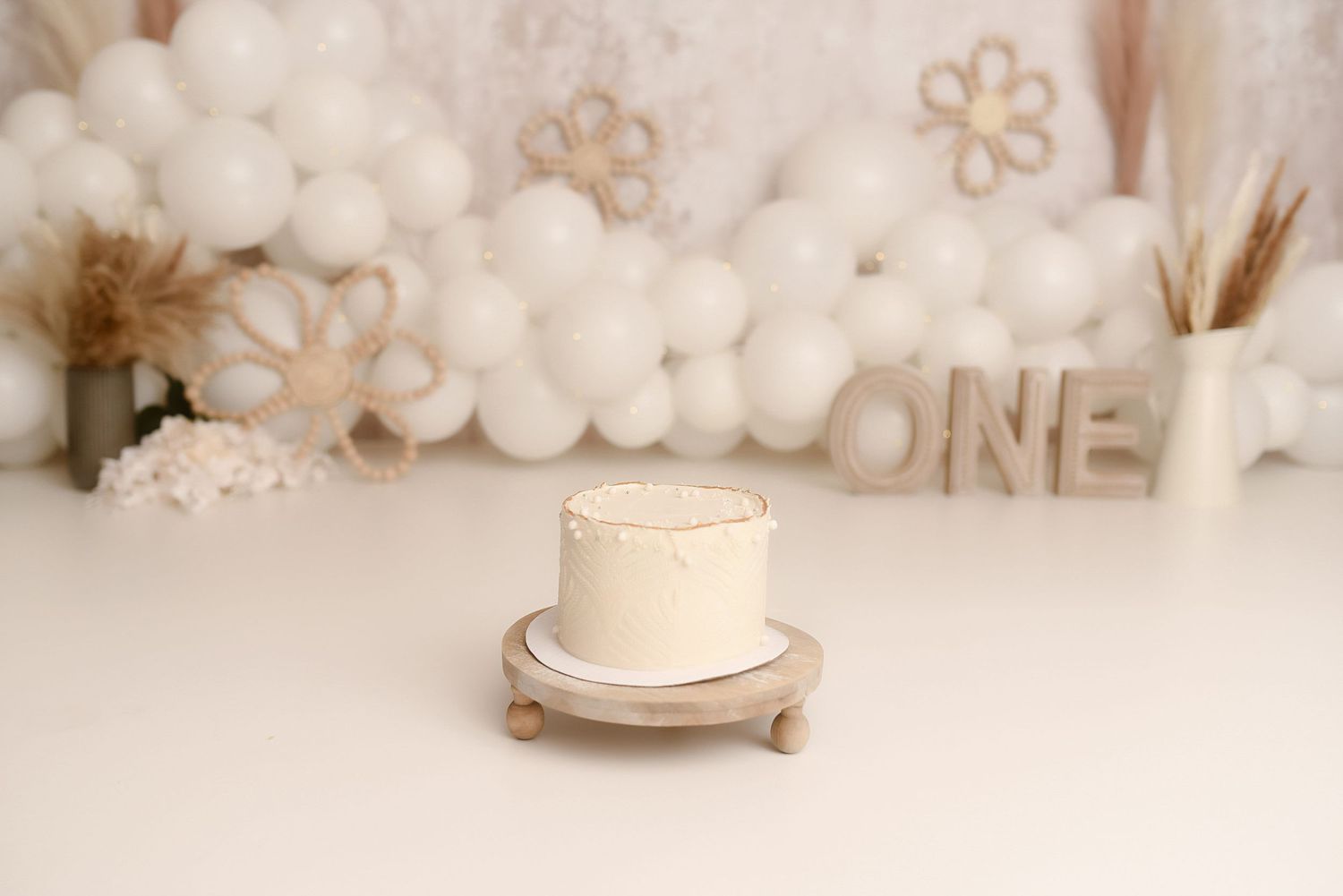 Simple white first birthday smash cake on wooden stand with boho balloon arch and ONE letters backdrop.