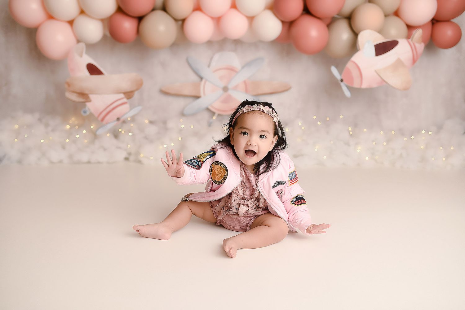 Baby girl in pink aviator jacket sitting before pink airplane and balloon first birthday backdrop.