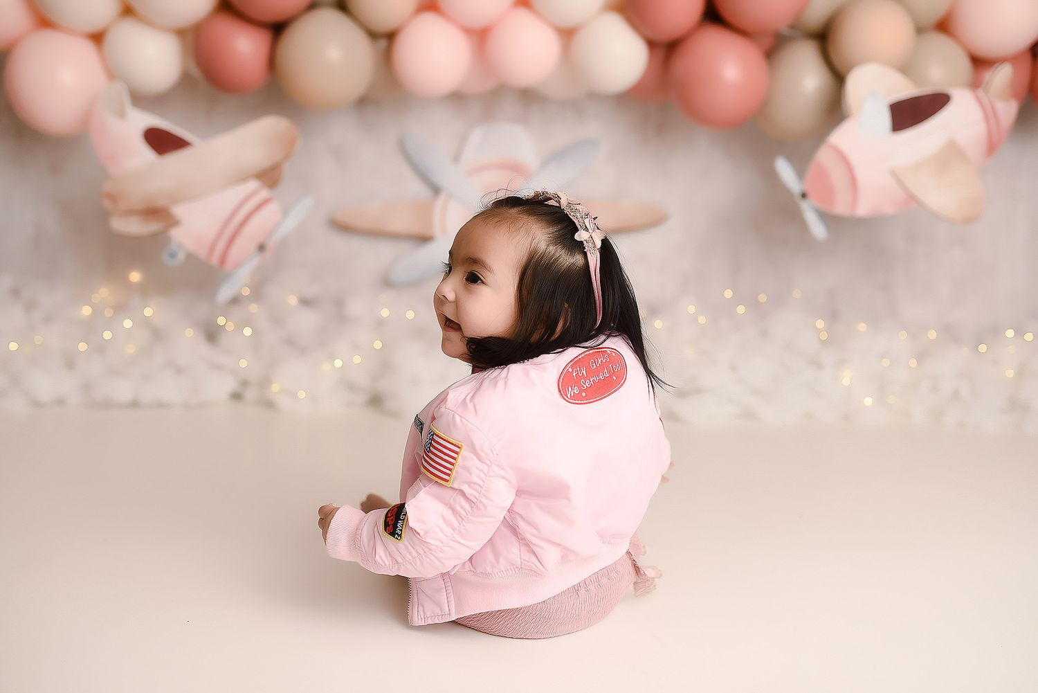 Baby girl in pink aviator jacket looking back during airplane themed first birthday photo session.
