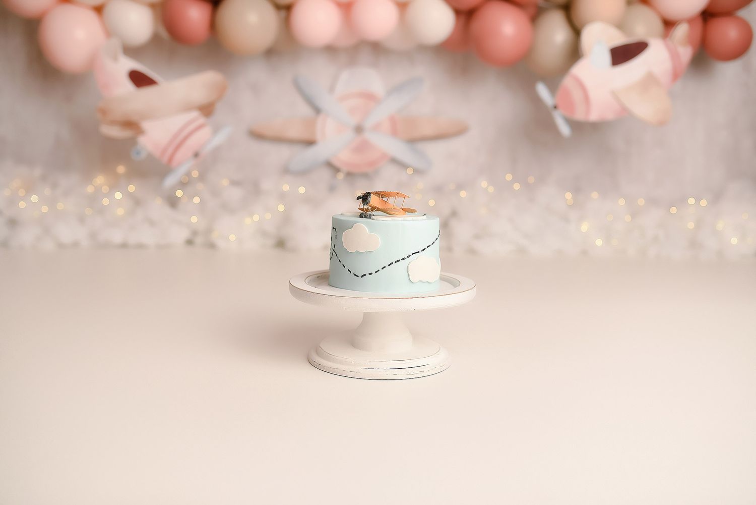 Light blue smash cake on white pedestal with airplane and balloon backdrop for first birthday.