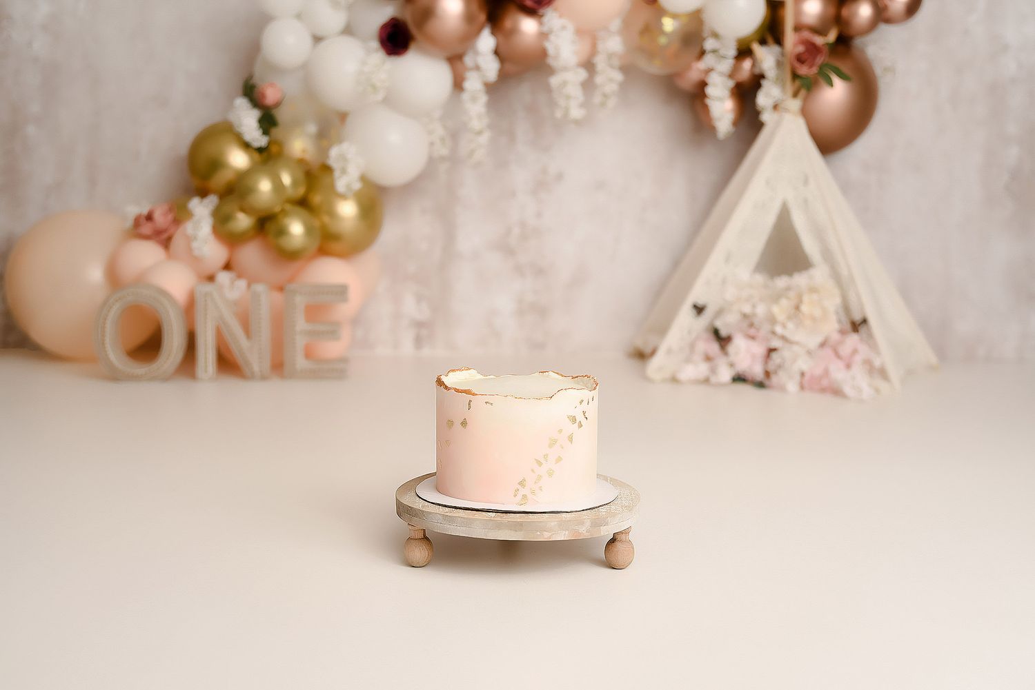 Pink smash cake on silver stand with ONE letters and balloon garland backdrop for first birthday.