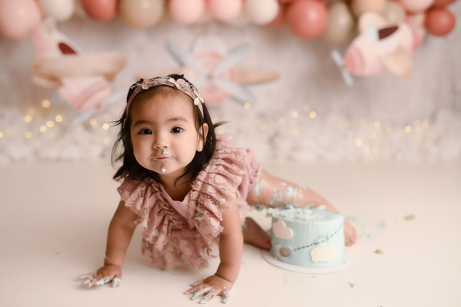Baby girl in pink dress crawling toward smashed blue cake during first birthday photoshoot.