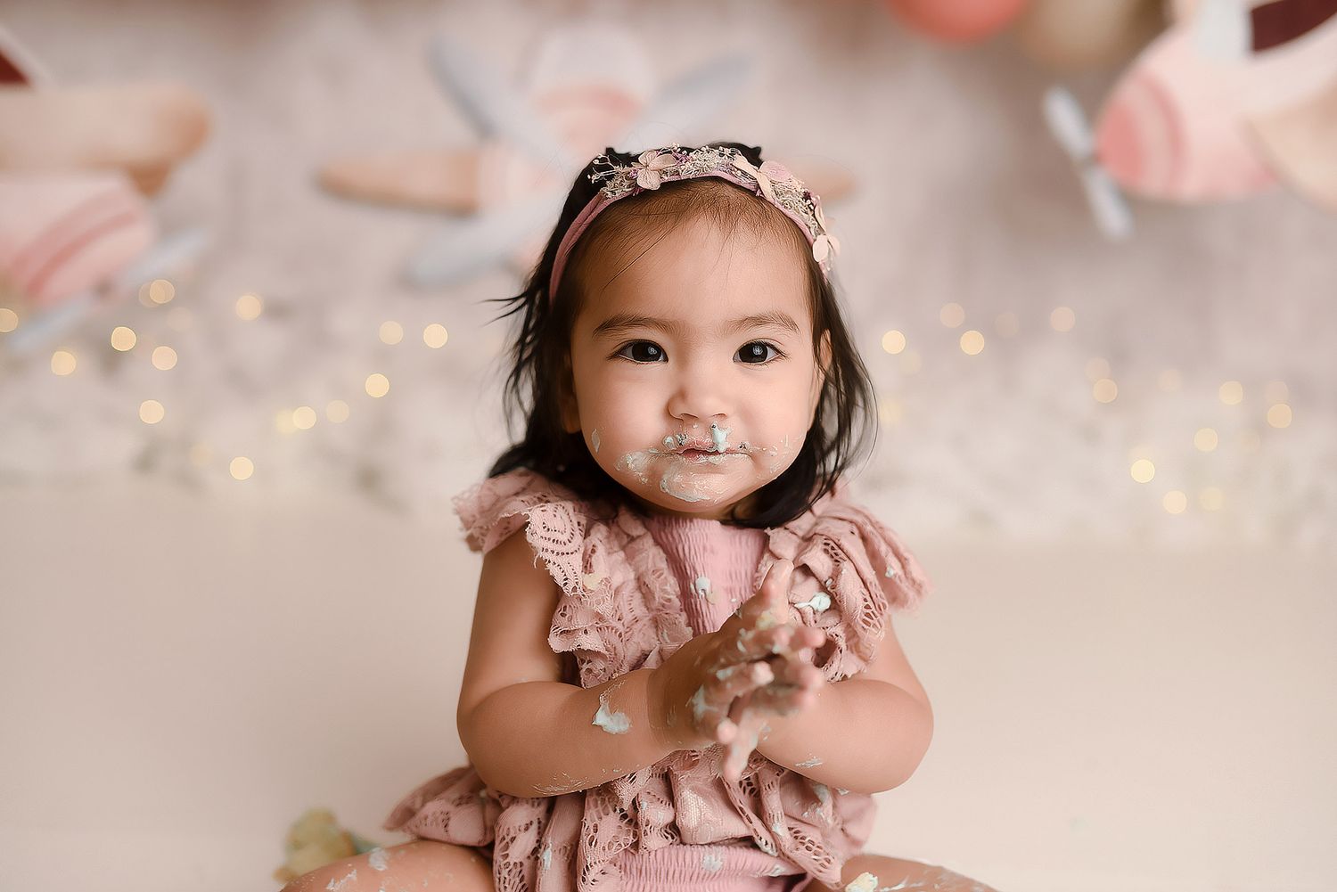 Smiling baby girl in pink dress with frosting on face during cake smash photoshoot.