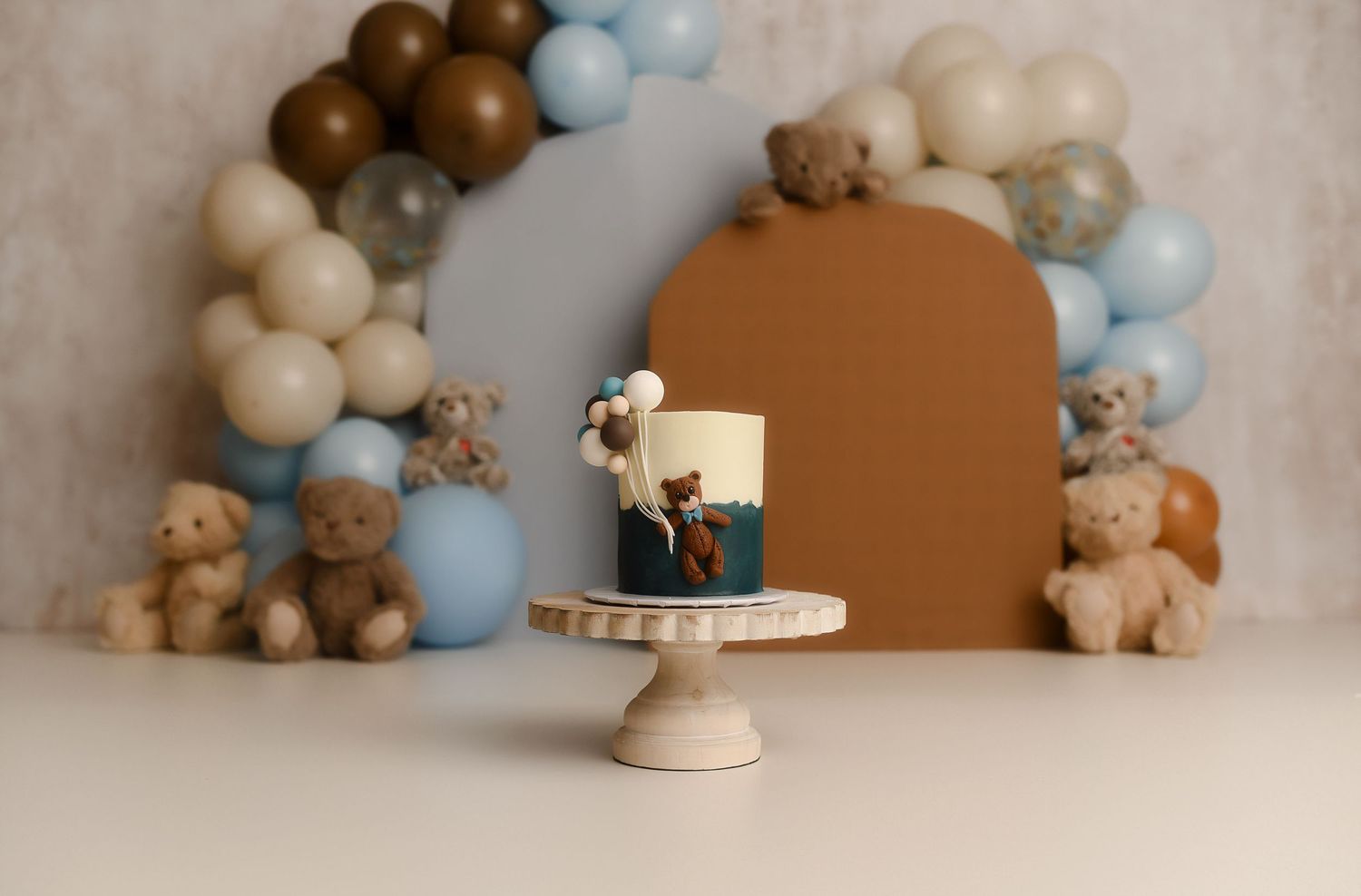 Teddy bear first birthday smash cake setup with blue and brown balloon arch, stuffed bears, and decorated cake on white stand.