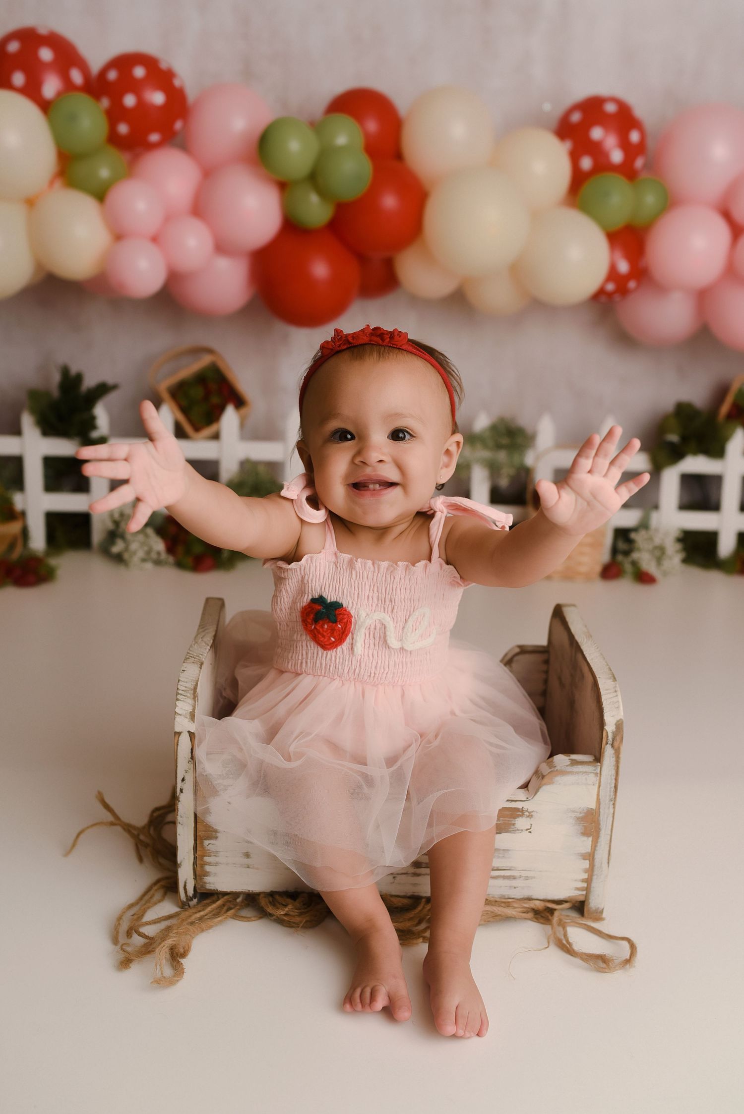 Smiling baby girl in pink tutu dress sitting in white wooden box with colorful balloon garland backdrop.