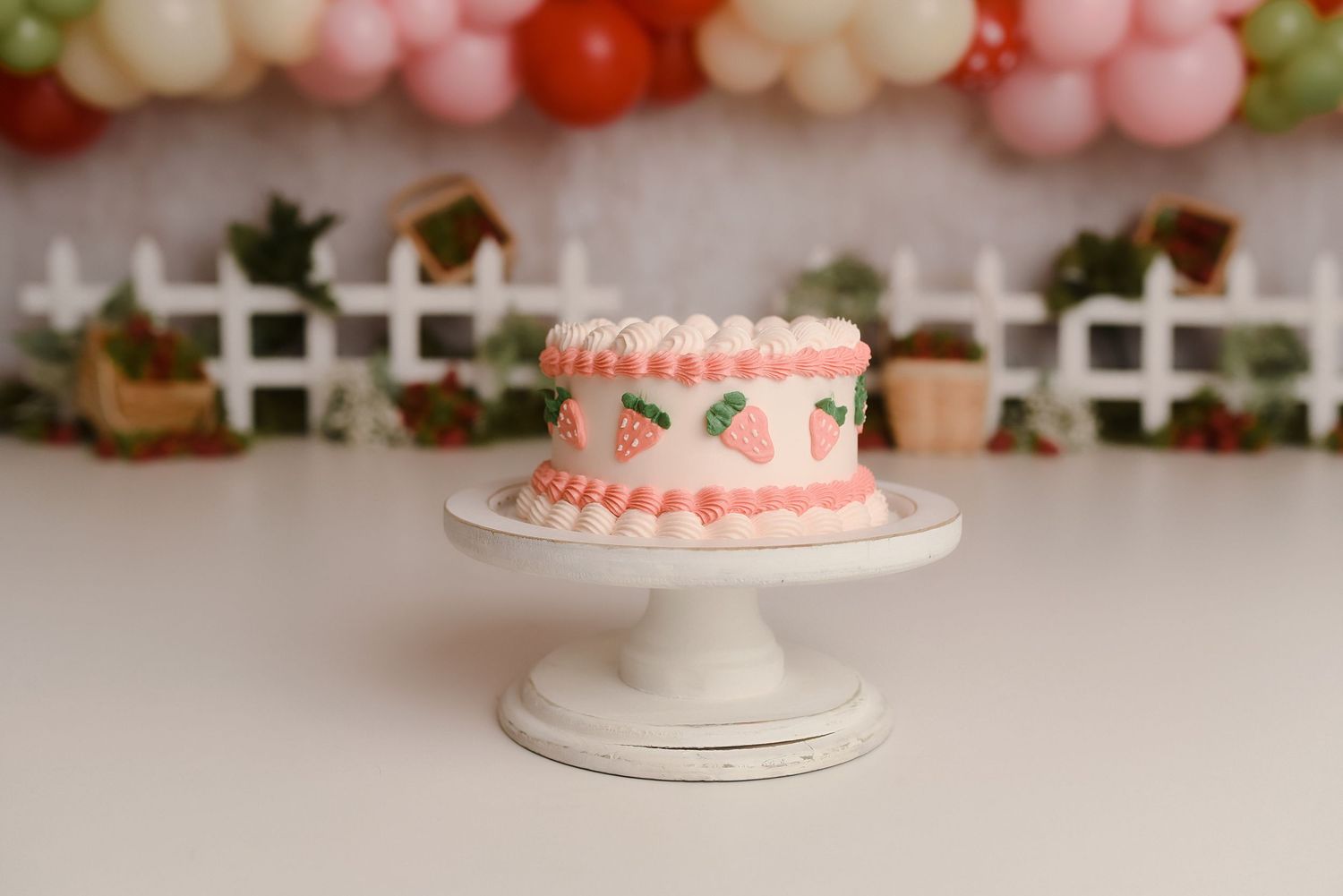 Pink birthday cake with strawberry decorations on white stand, balloon garland backdrop.
