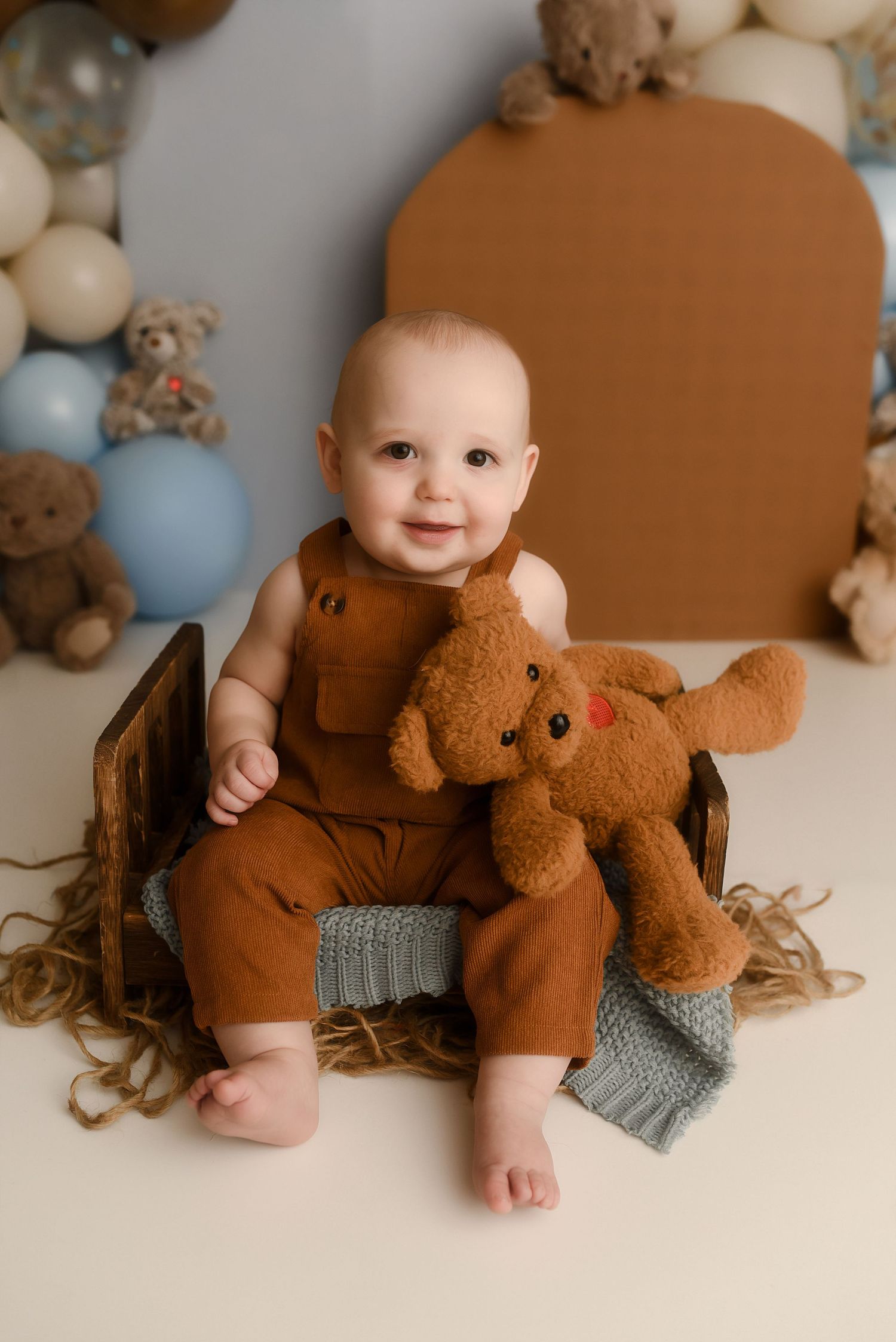 Smiling baby in brown overalls sitting in wooden crate with teddy bear, surrounded by balloons and stuffed animals.