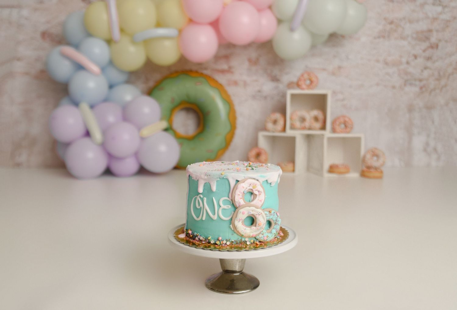 Teal first birthday cake reading ONE with donut-themed balloon garland and pastel backdrop.