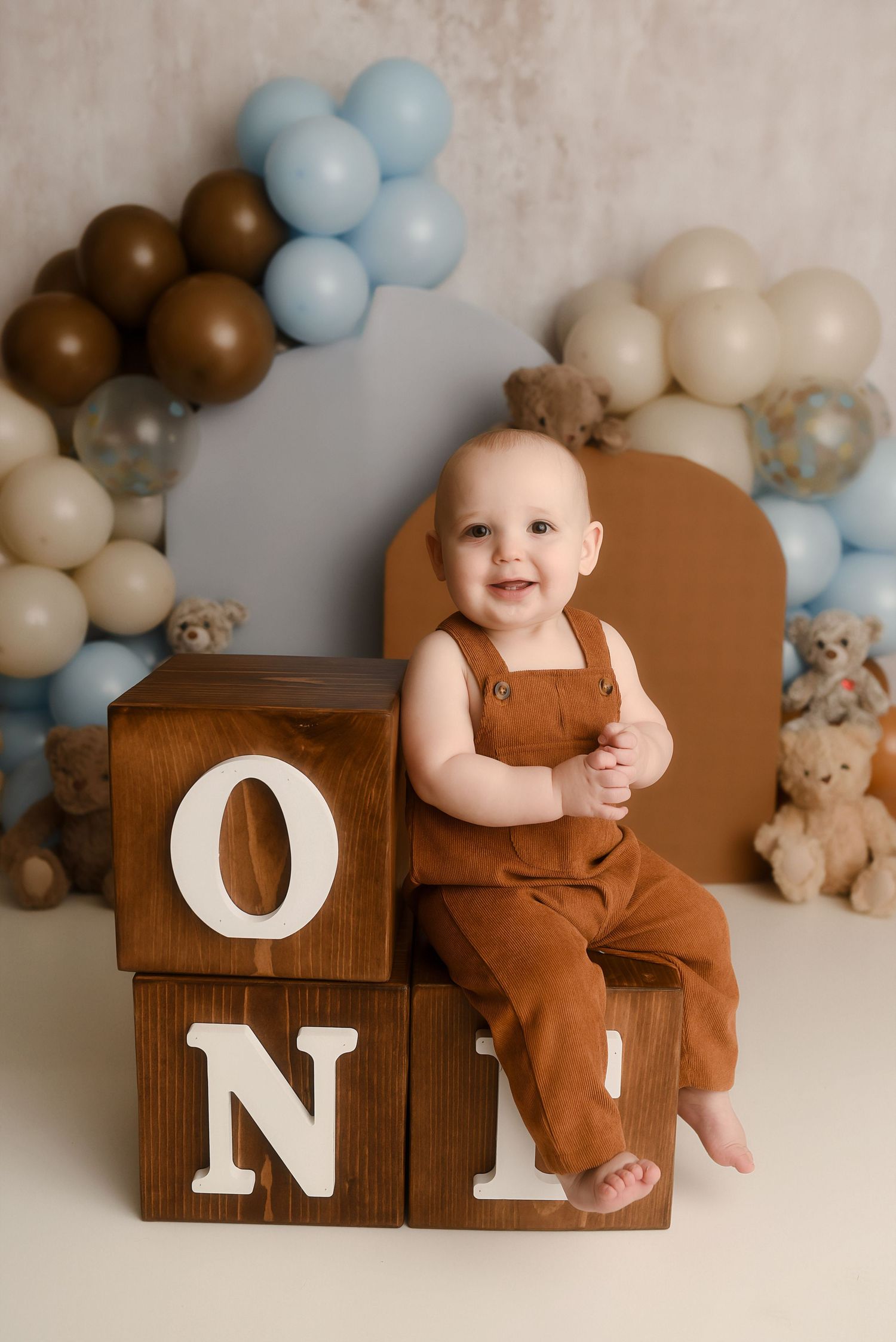 Baby boy in brown overalls sits on wooden ONE blocks at his first birthday party with blue and brown balloon decorations.