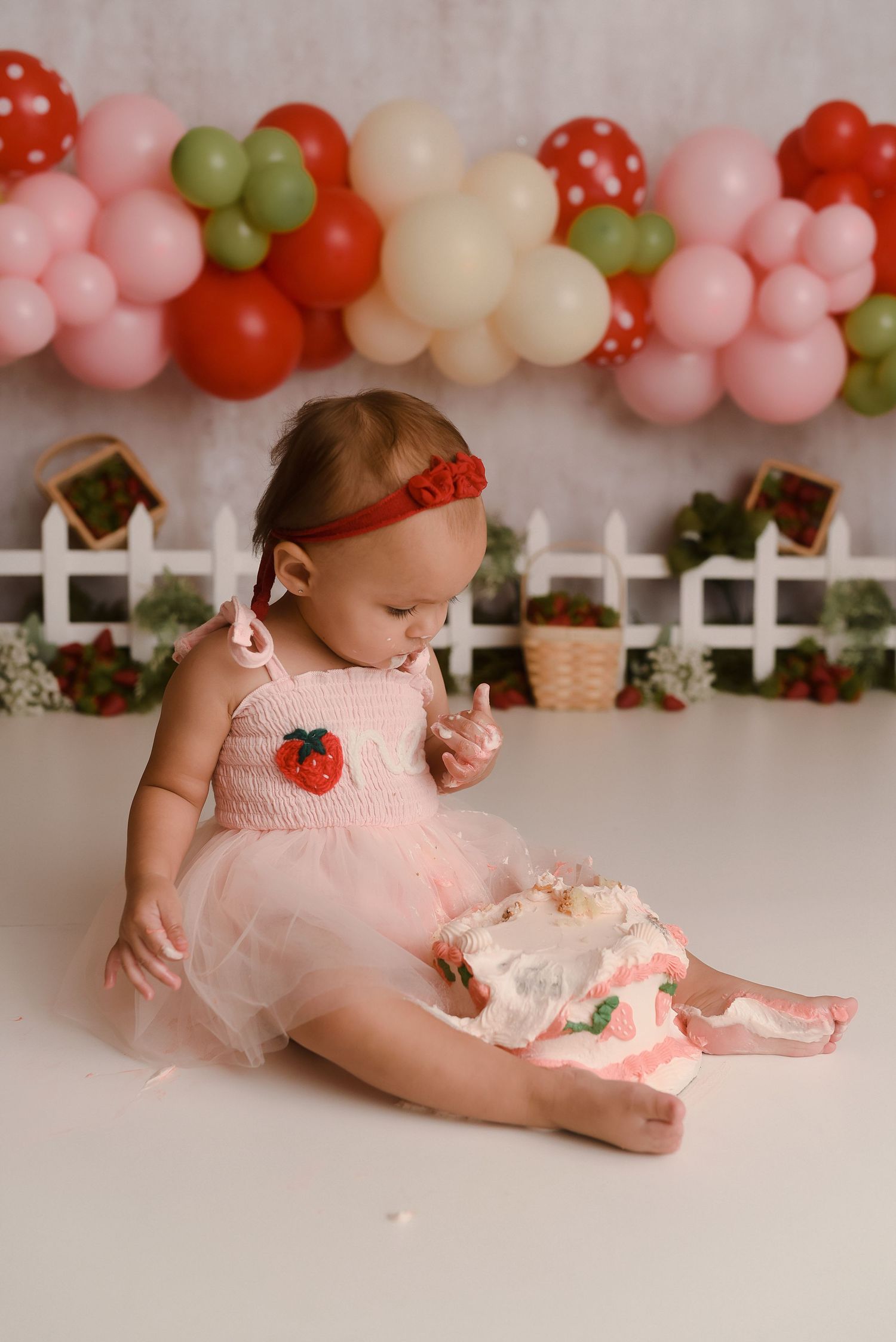 Baby girl in pink tutu dress smashing her first birthday strawberry cake, surrounded by colorful balloon garland backdrop.