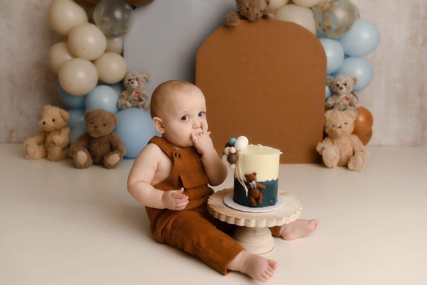 Baby in brown romper sits beside a smash cake, surrounded by teddy bears and blue balloons at a first birthday photoshoot.