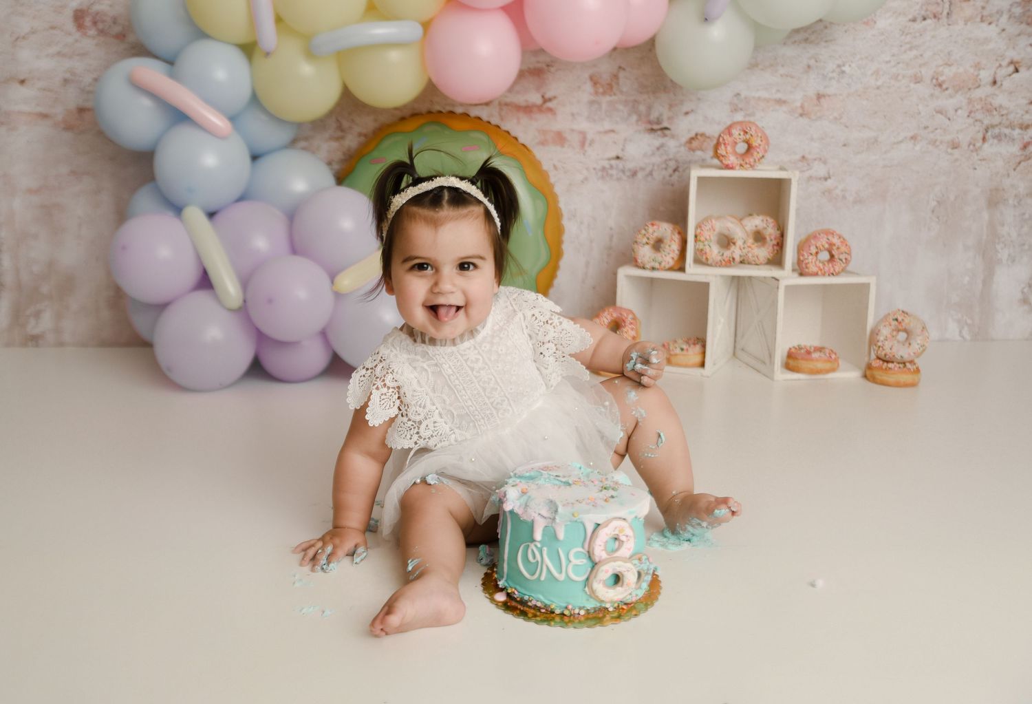 Baby girl smiling with teal first birthday cake smash in donut and pastel balloon themed studio.