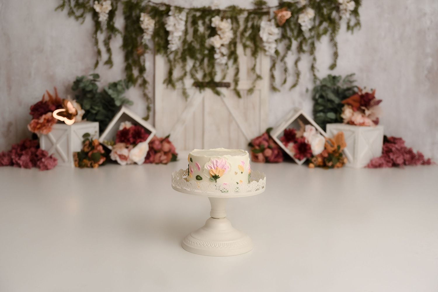 White cake with floral decorations on a pedestal stand, surrounded by flowers and greenery backdrop.