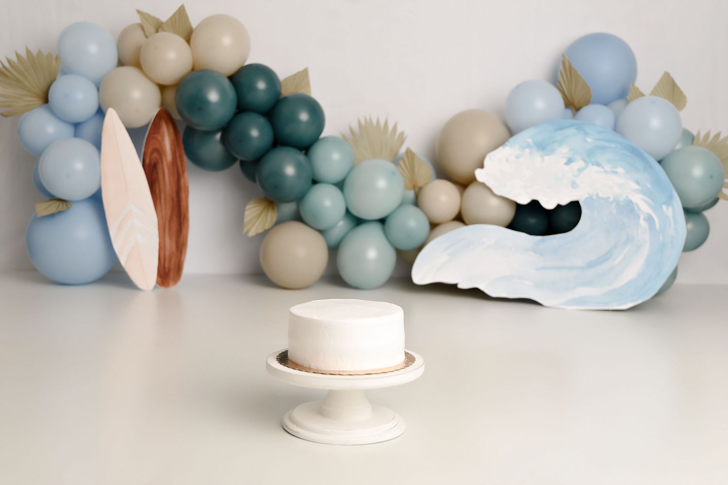 White first birthday cake on pedestal with ocean wave prop and blue balloon garland backdrop.