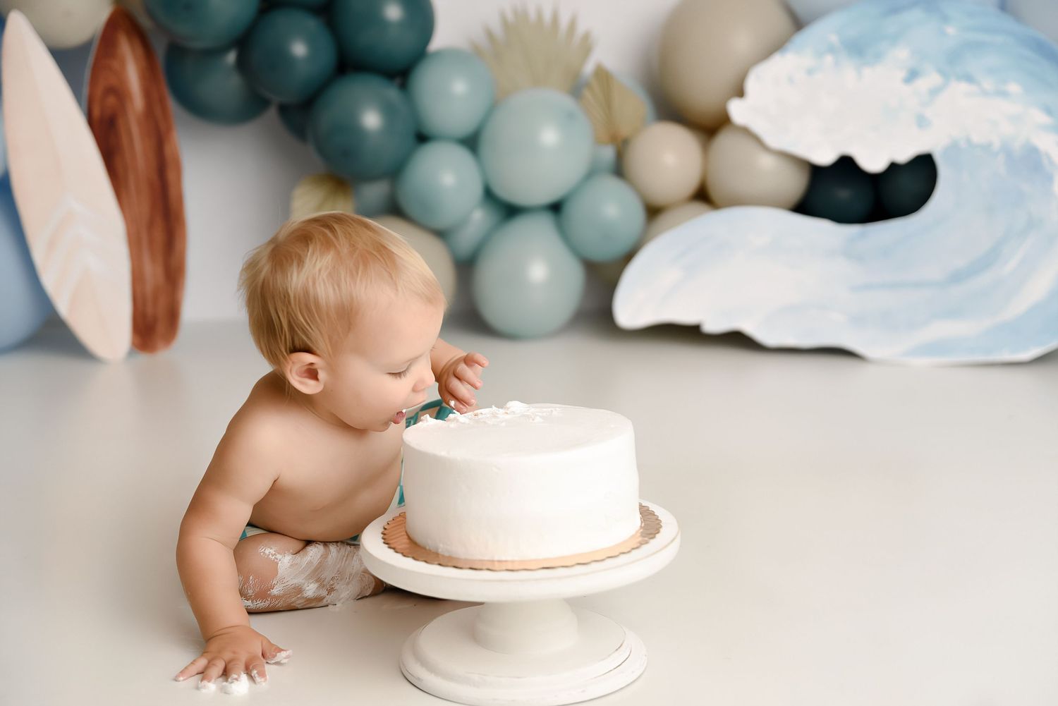 Toddler boy eating white first birthday cake with ocean wave and balloon garland backdrop.