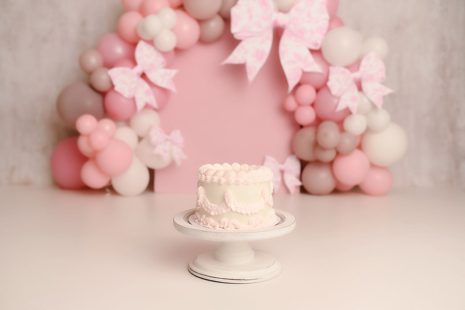 White ruffled first birthday smash cake on stand with pink balloon and bow arch backdrop.