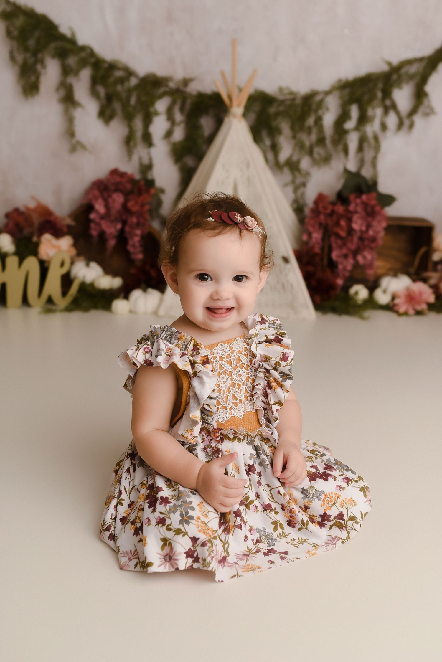 Smiling baby girl in floral dress sits on white floor with teepee and flower backdrop for first birthday photo session.