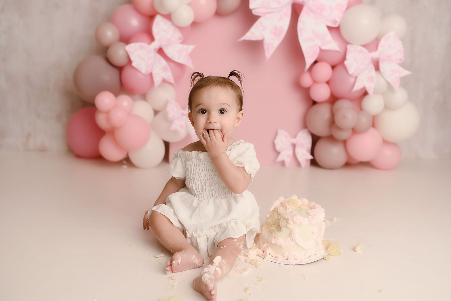 Baby girl in white dress smashing first birthday cake with pink balloon and bow arch backdrop.