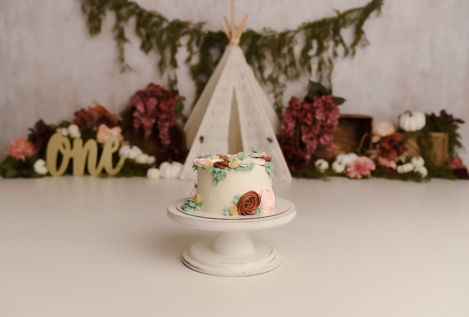 A floral smash cake on a white pedestal stand with a boho teepee and flower backdrop for a first birthday.