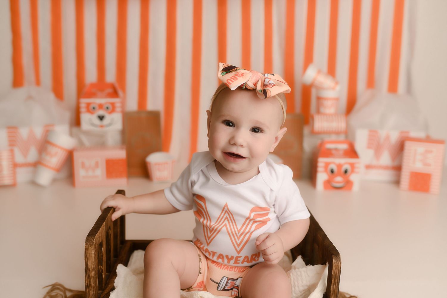 Adorable baby girl in Whataburger onesie sitting in wooden basket with orange and white themed party decorations.