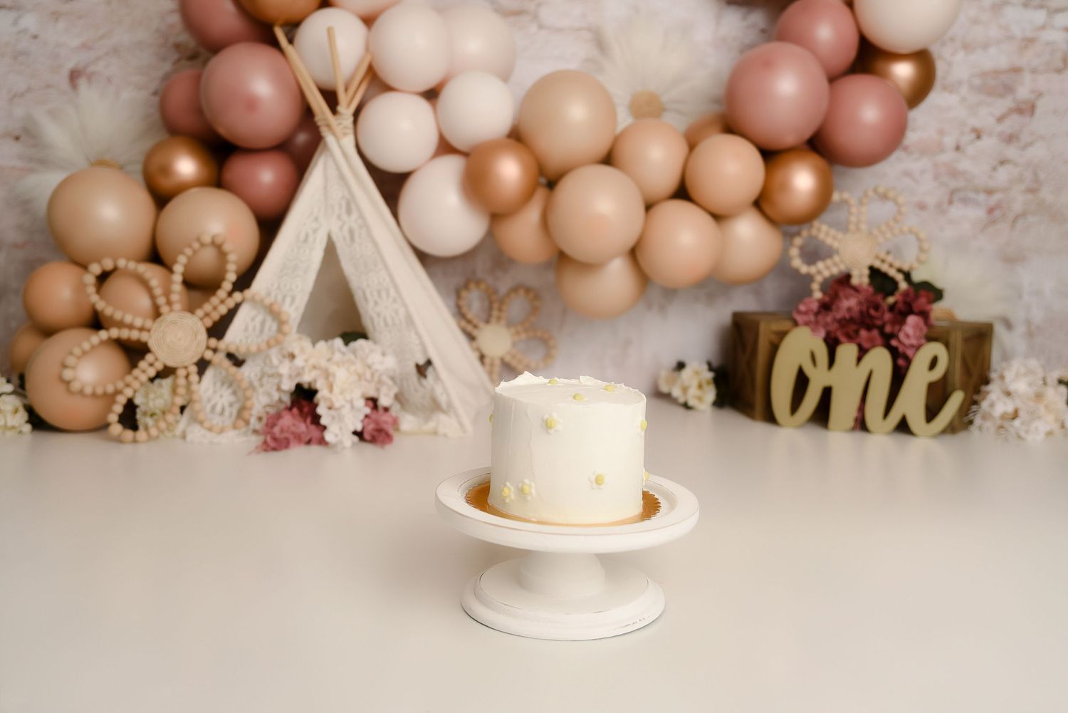 White first birthday smash cake on pedestal with boho balloon garland and teepee backdrop.