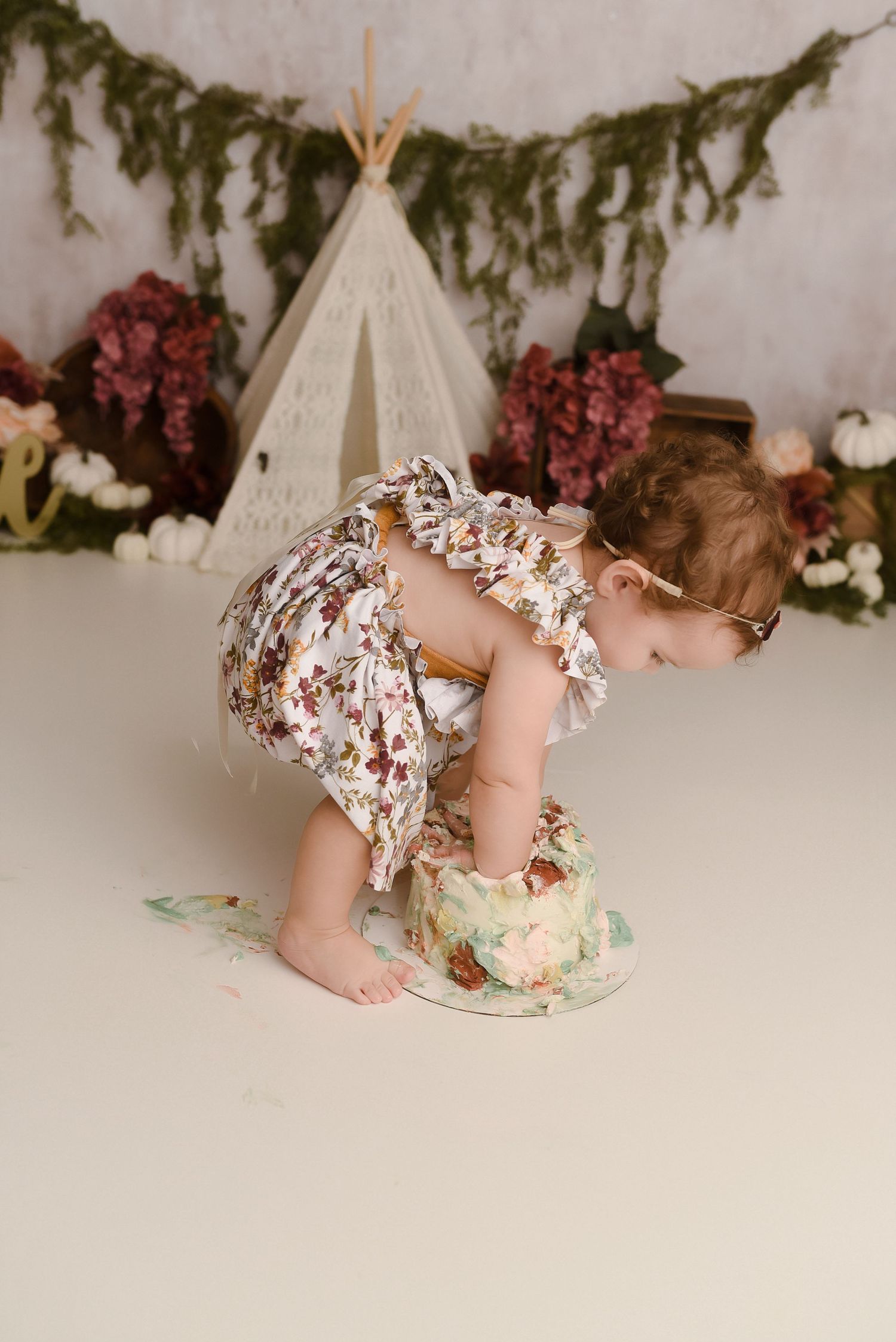 A baby in a floral romper smashes a first birthday cake in front of a boho teepee backdrop with greenery.
