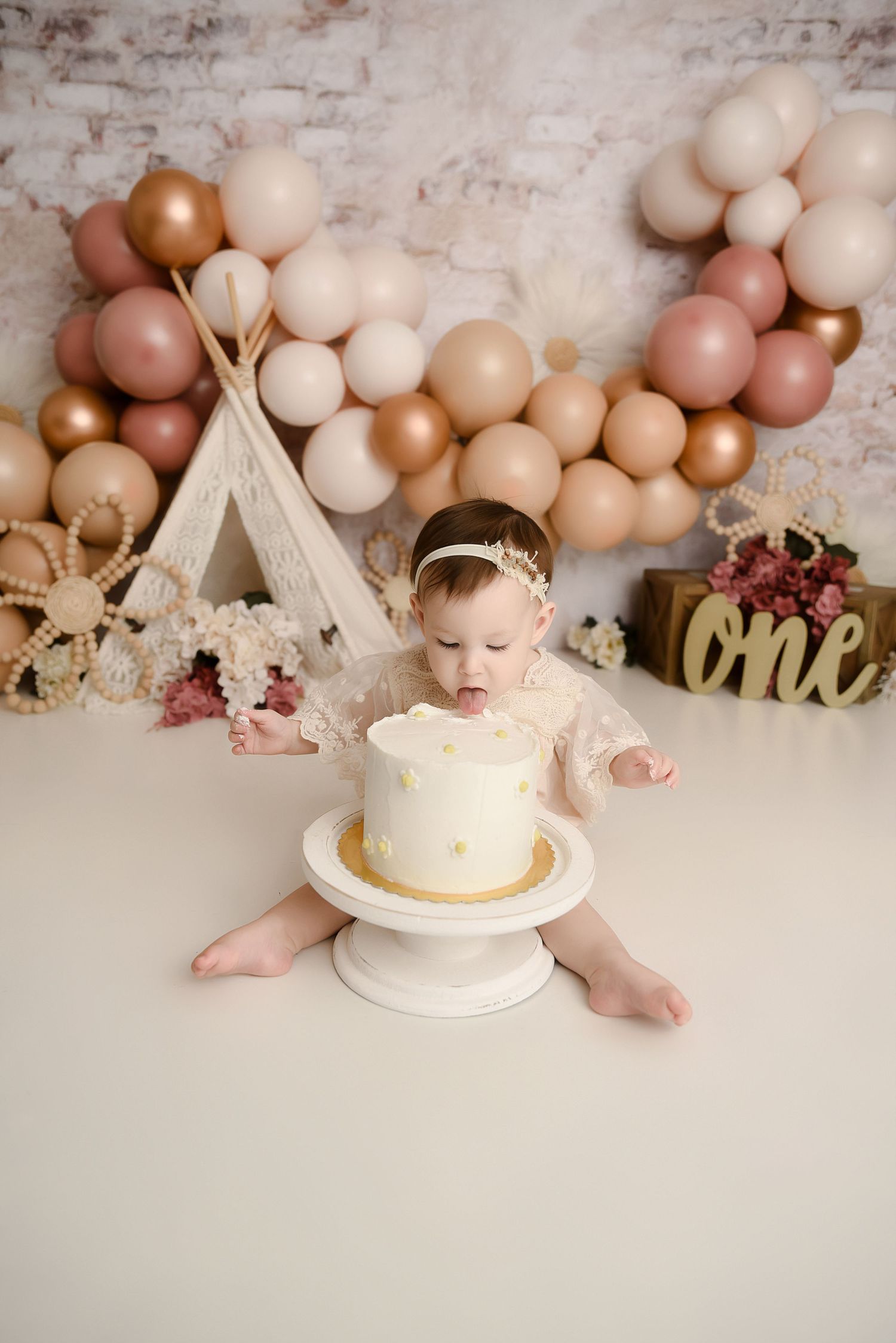 Baby girl in lace outfit touching first birthday smash cake with boho balloon backdrop.