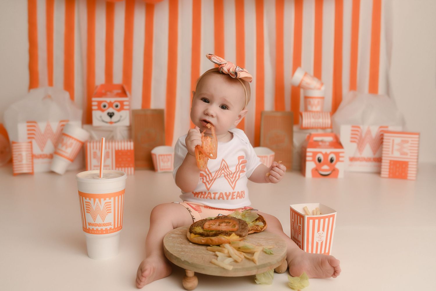 Baby in Whataburger onesie sits surrounded by Whataburger themed decorations, food, and drinks for a first birthday cake smash.