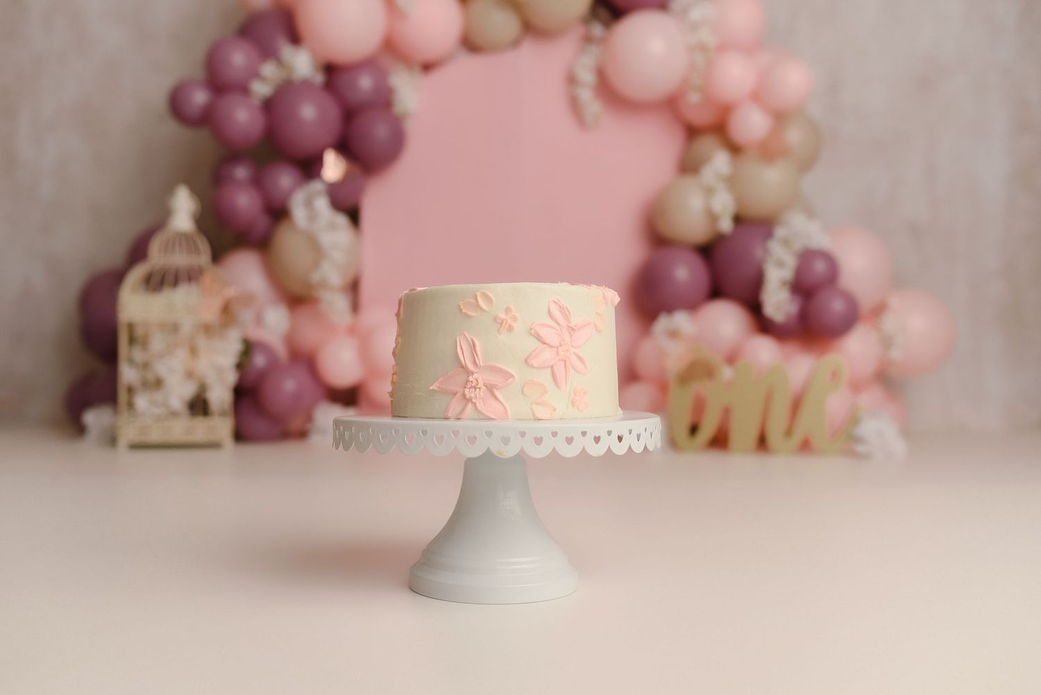 A white cake with pink floral details on a cake stand, surrounded by pink and mauve balloon arch backdrop.