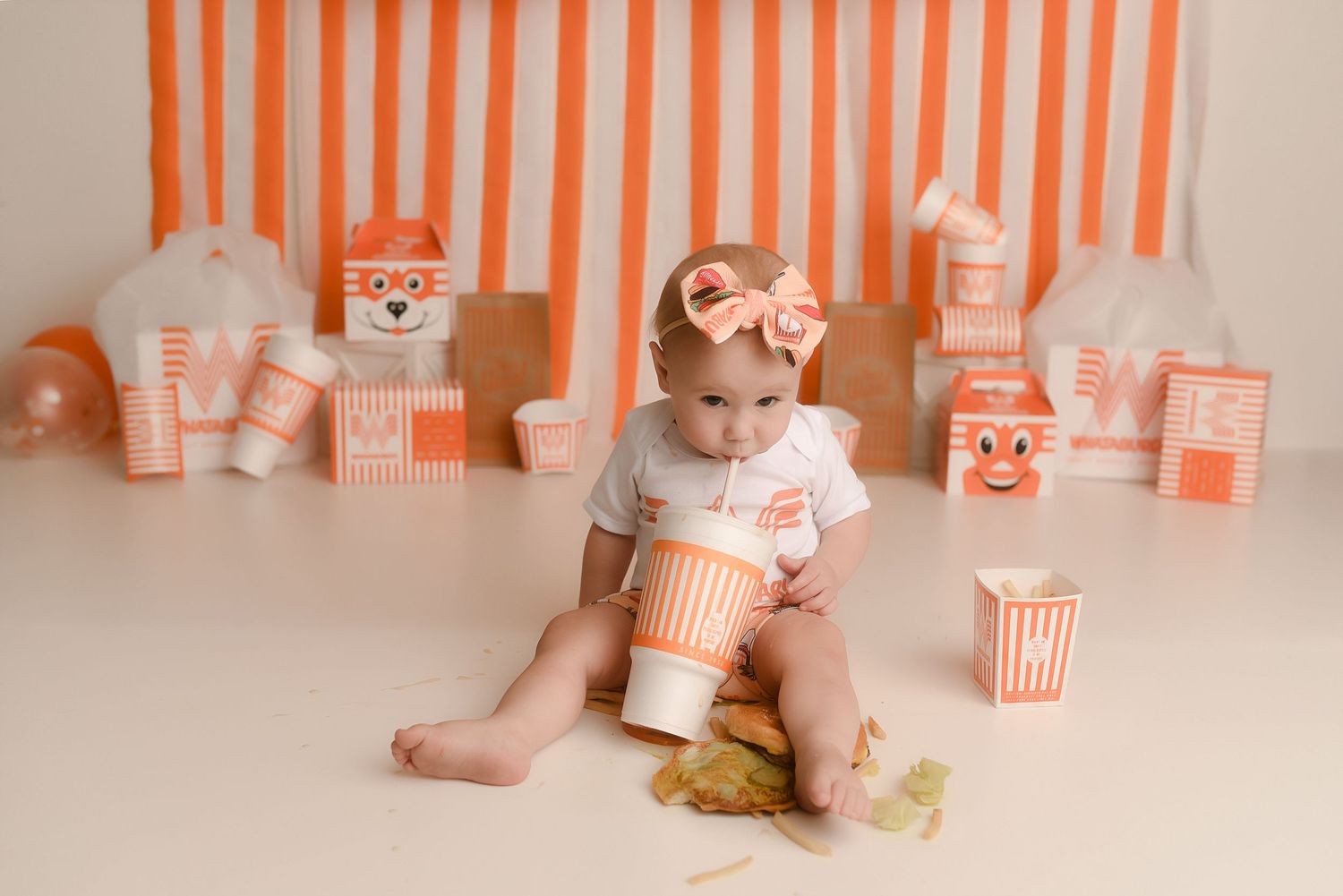 Baby girl in white onesie with bow headband sits among orange striped backdrop and festive blocks during cake smash photo session.