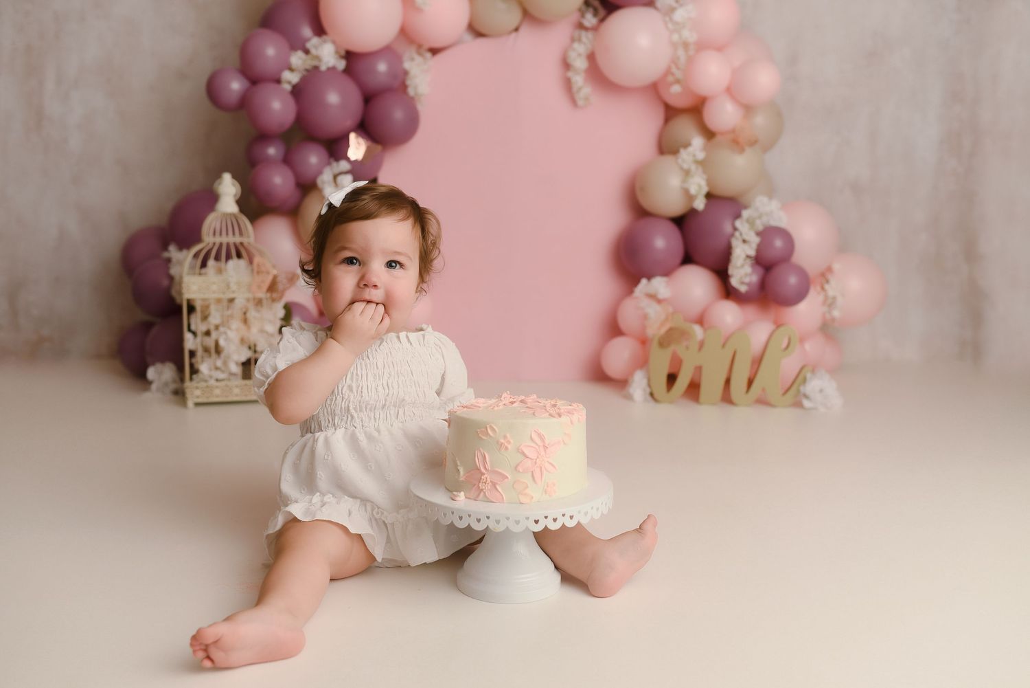 Baby girl in white romper smashes first birthday cake surrounded by pink balloon arch and floral decorations.