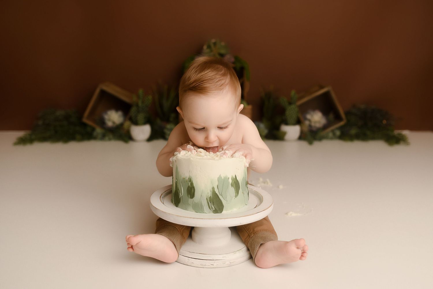 Baby boy in brown pants smashes green leaf cake at nature-themed first birthday photo session.