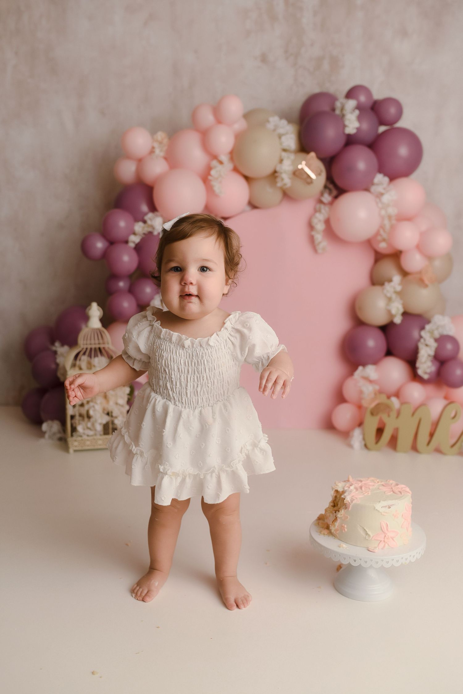 Baby girl in white dress stands by first birthday cake with pink and mauve balloon arch backdrop.