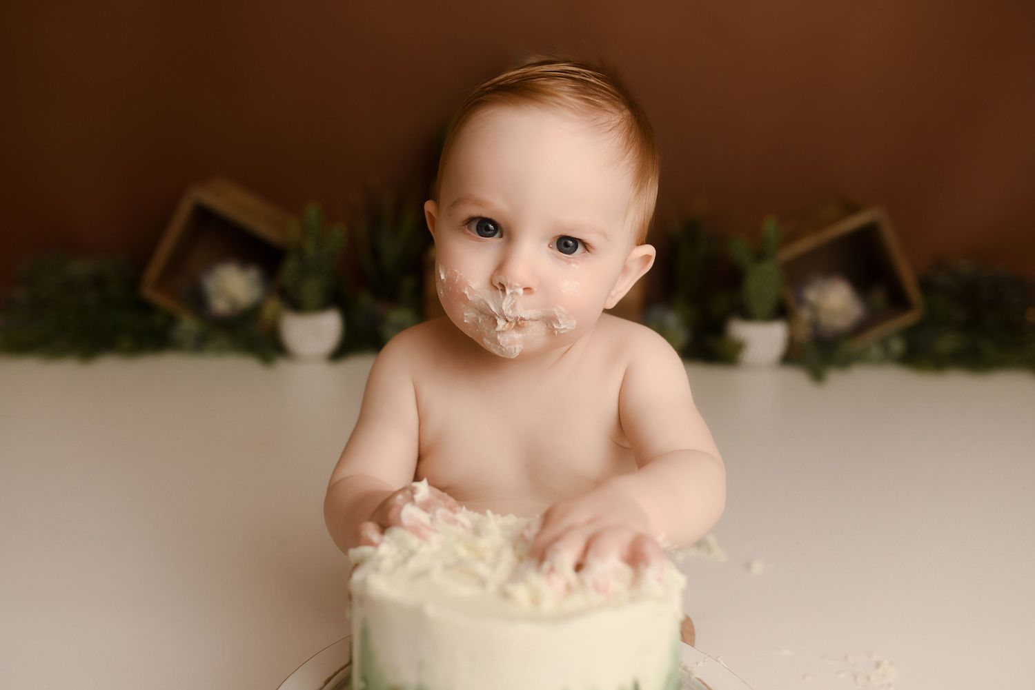 Close-up of baby boy with frosting on face touching green and white first birthday cake.