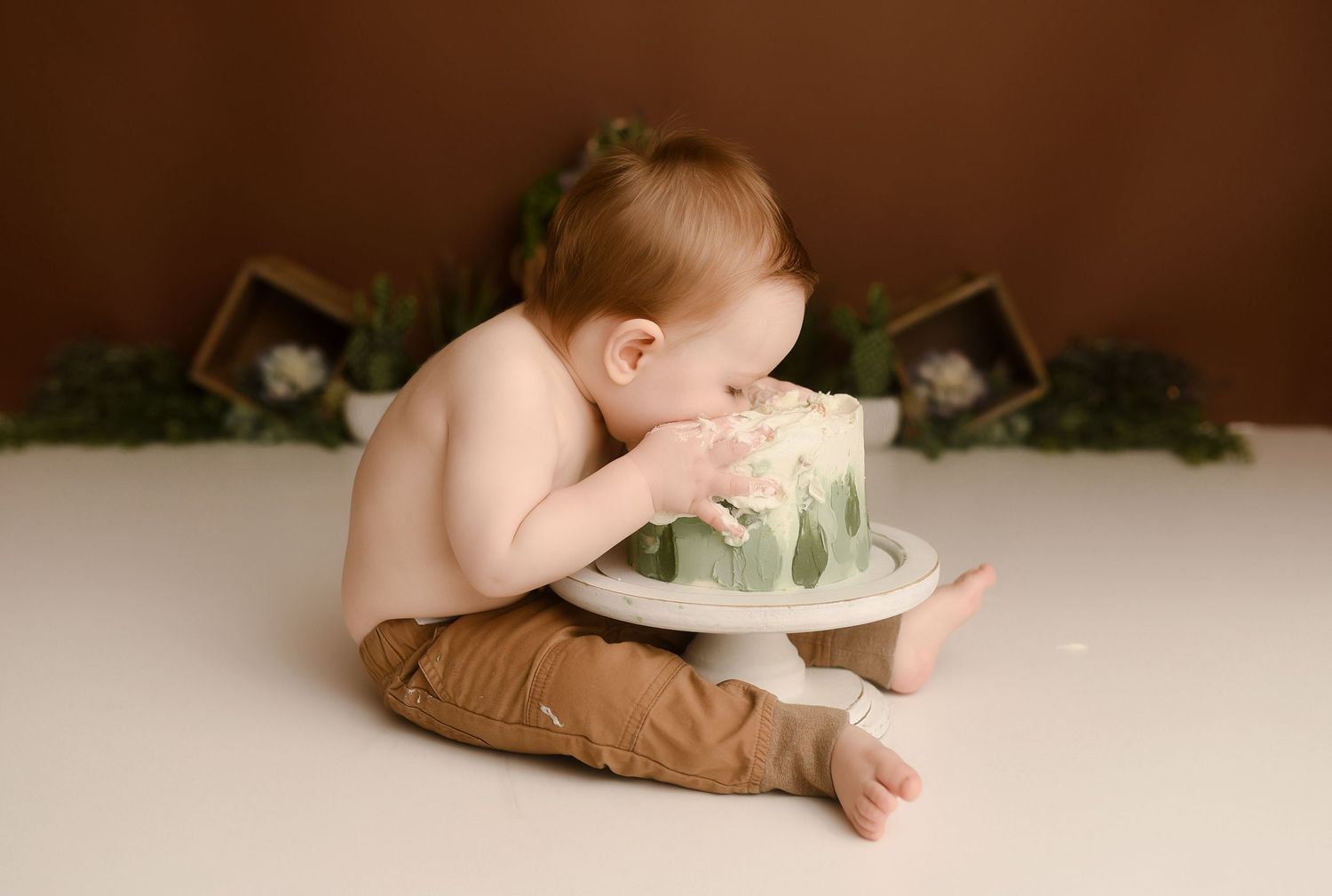 Baby boy in brown pants leans into green leaf first birthday cake during cake smash session.