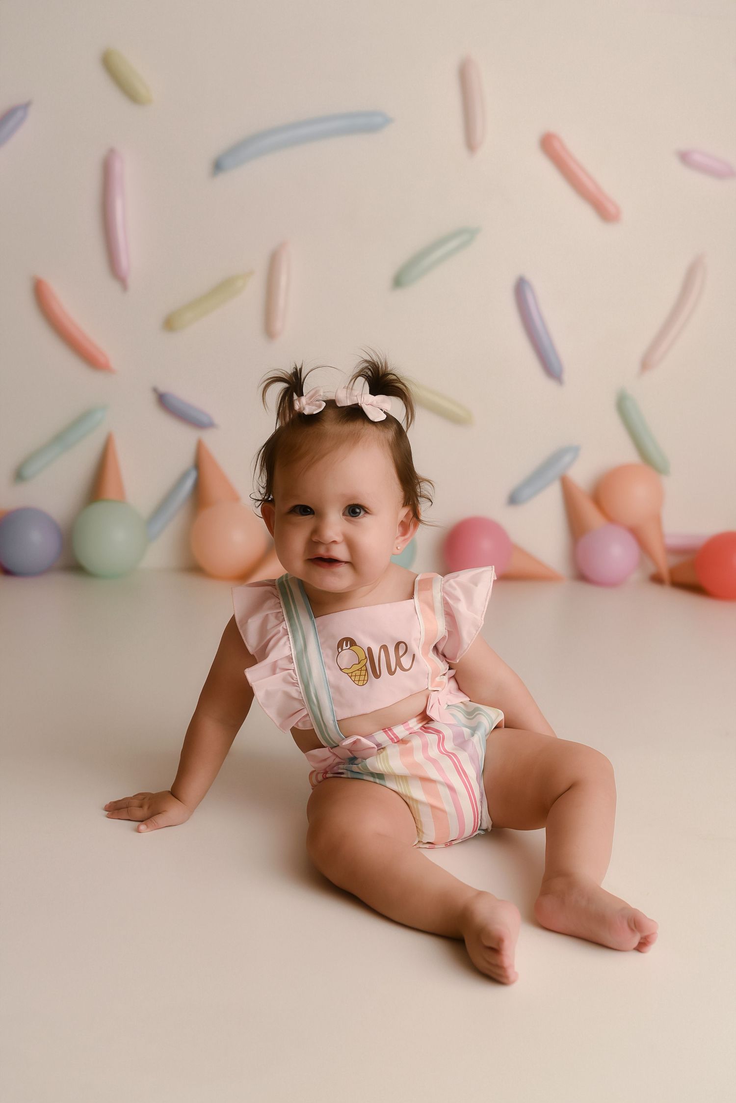 A baby girl in a pink striped romper with a bow headband sits on a white floor for her first birthday cake smash photo.