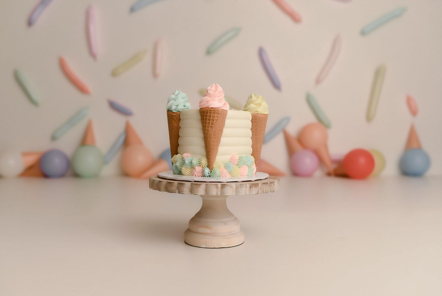 A whimsical ice cream cone cake on a wooden stand with pastel sprinkle backdrop and decorative cones.