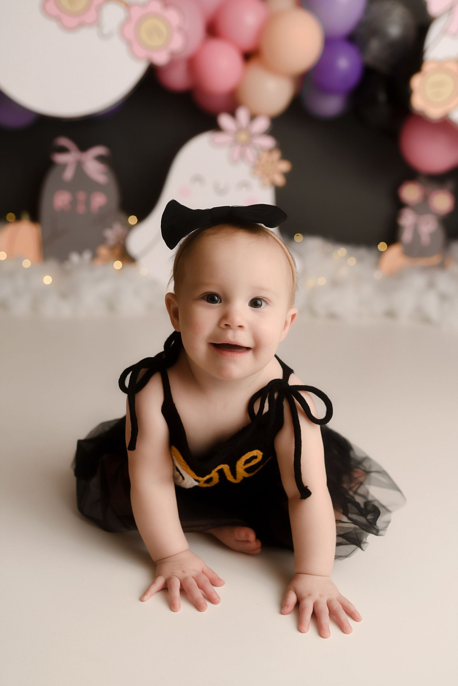 Smiling baby in black tutu outfit sitting in front of ghost Halloween themed birthday balloon backdrop.