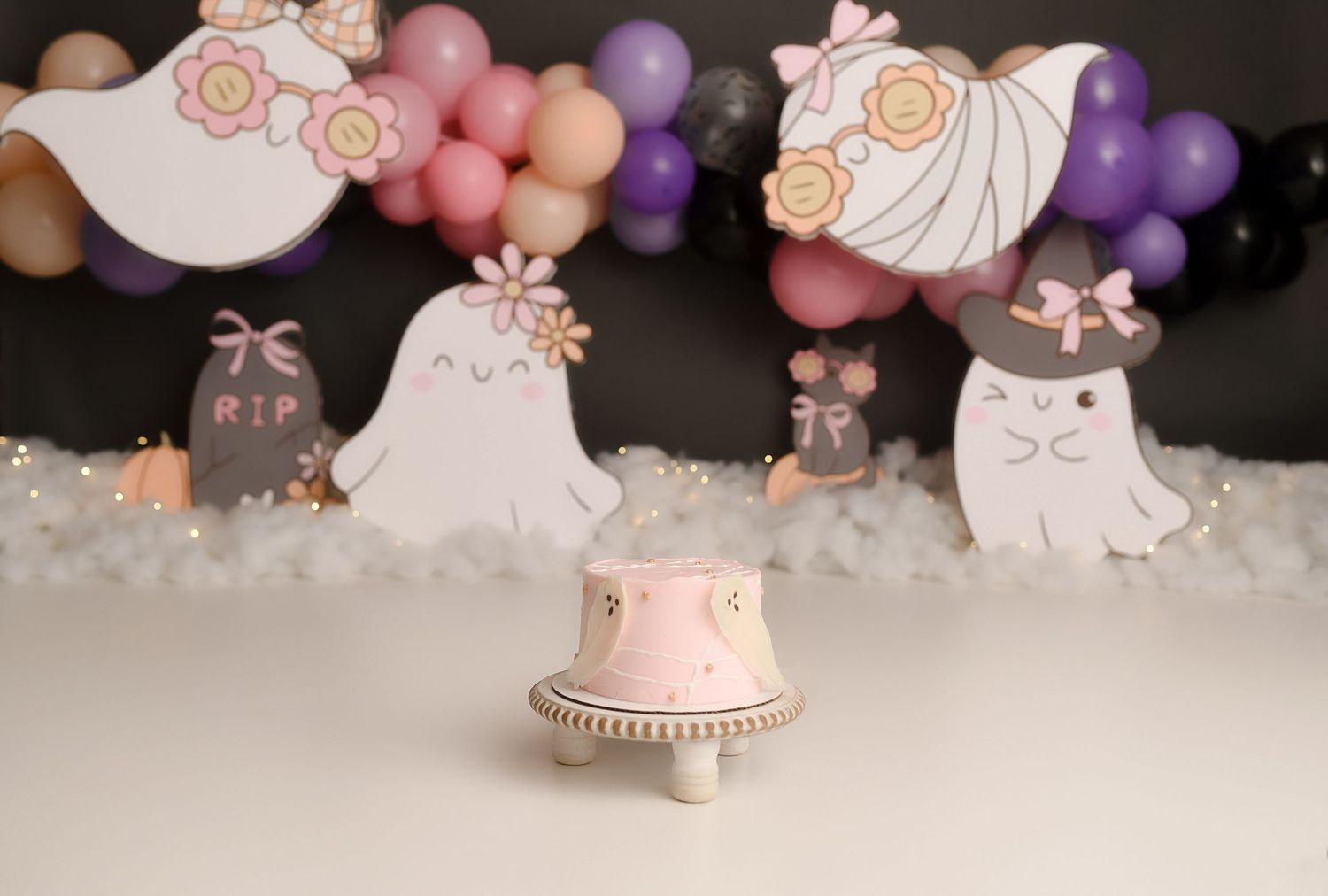 Cute ghost-themed first birthday cake smash setup with pink cake, RIP tombstone, and Halloween balloon arch.