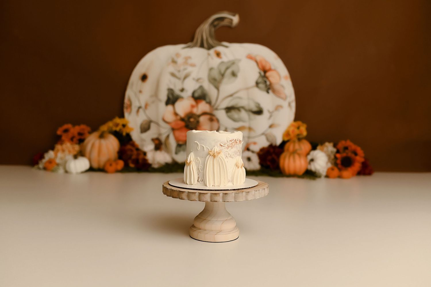 Small white drip cake on wooden stand with floral pumpkin and autumn flowers on brown background.