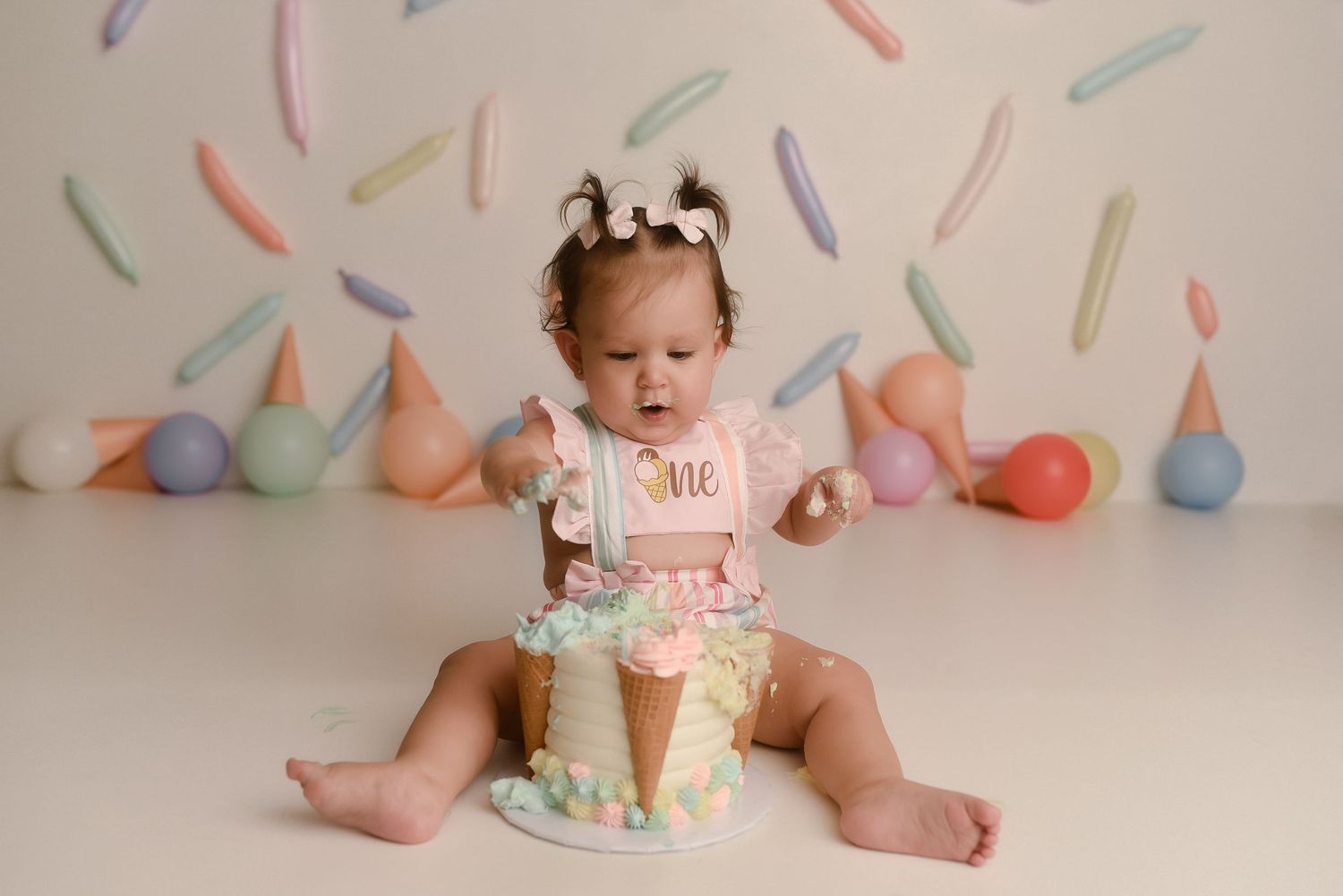 Baby girl in pink outfit smashes first birthday cake surrounded by pastel balloons and colorful sprinkle backdrop.