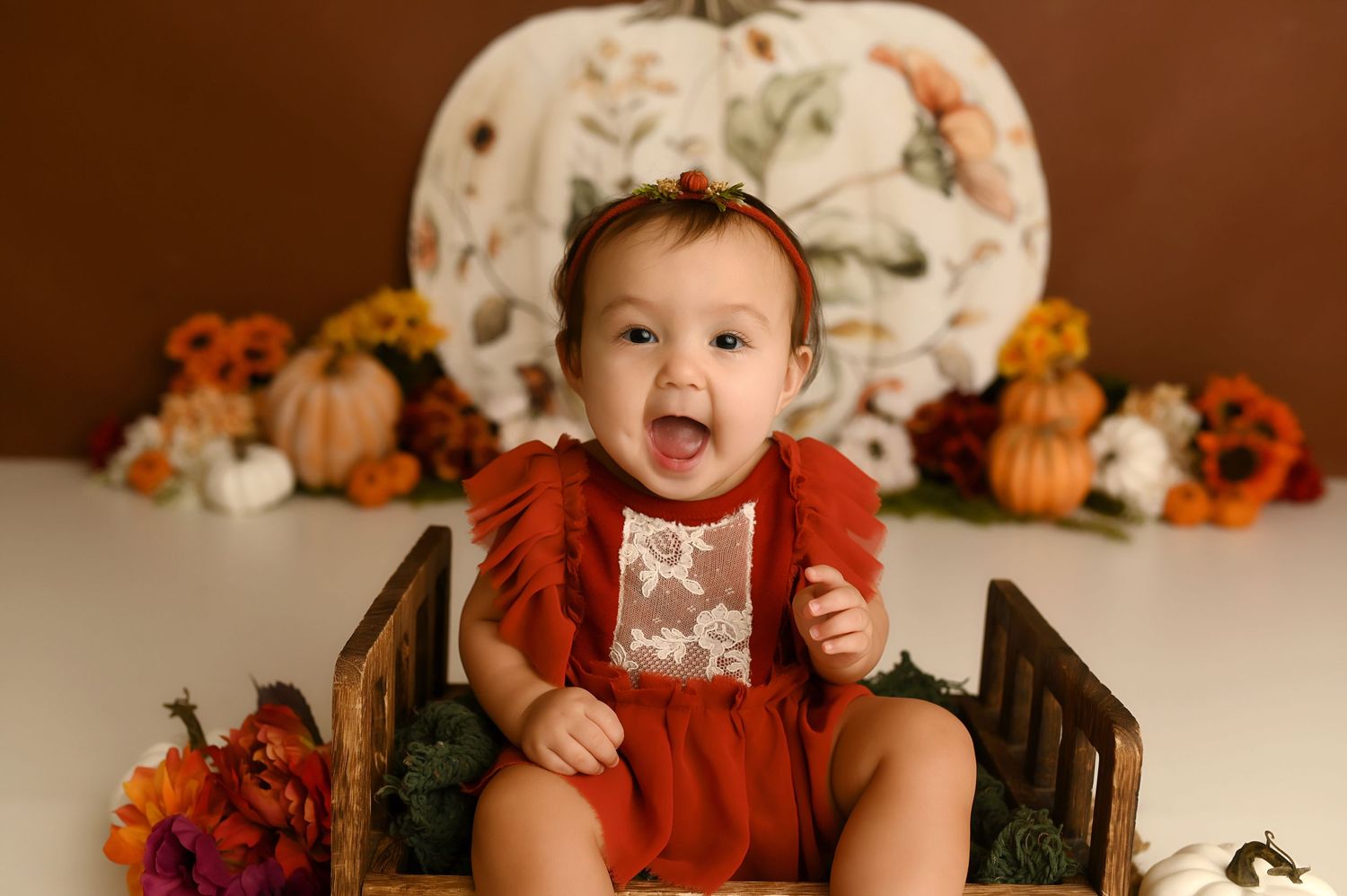 Smiling baby in red romper sits in wooden crate with fall pumpkins and flowers in autumn photo session.