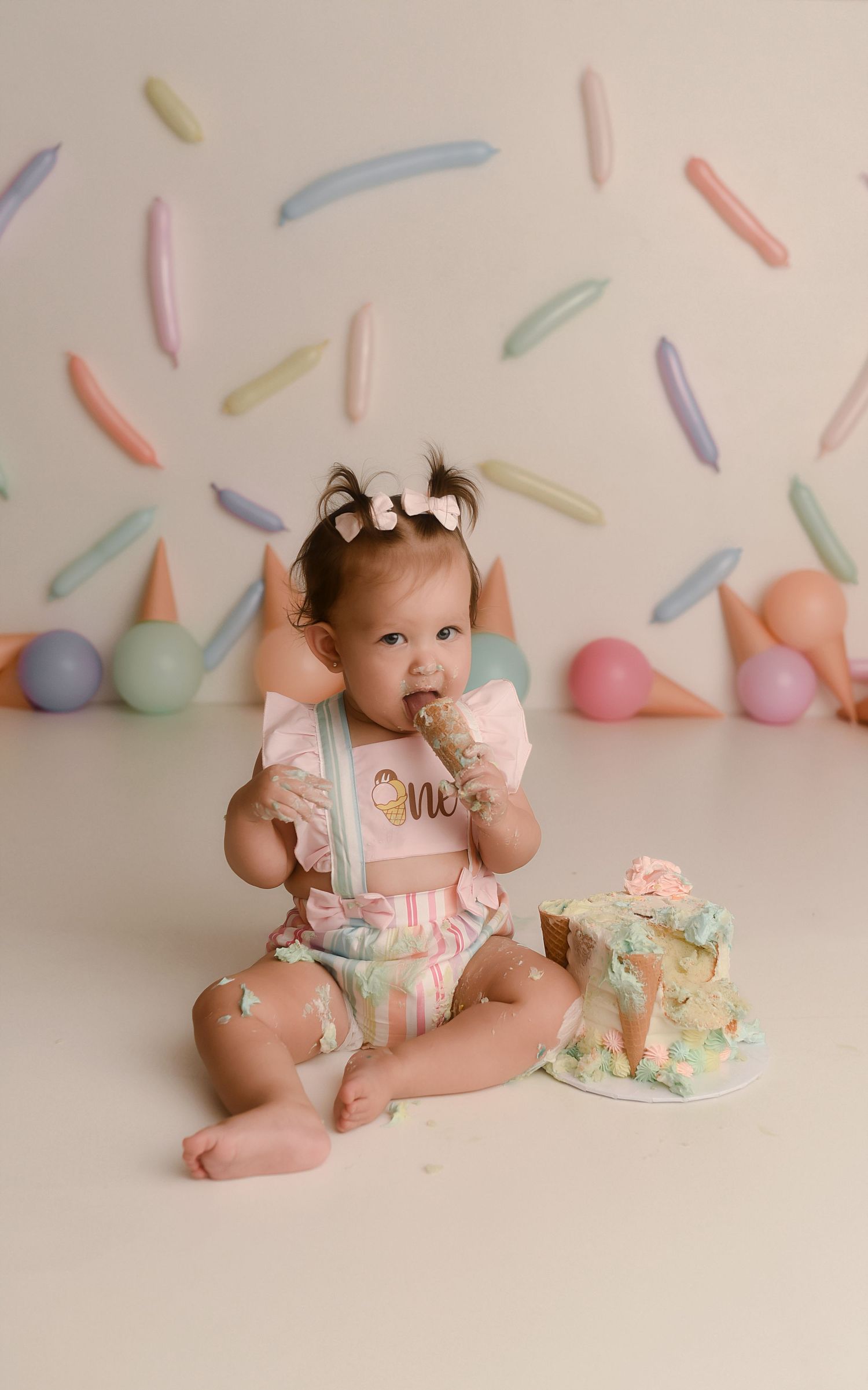Baby girl in pink outfit smashes first birthday cake during colorful sprinkle-themed cake smash photo session.