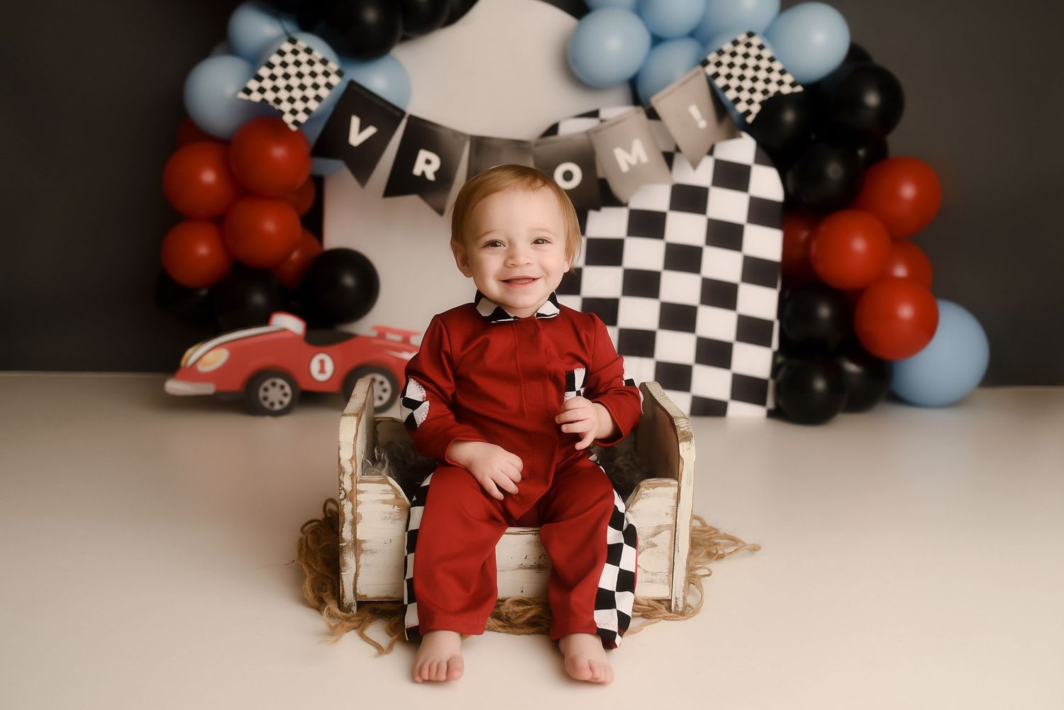 Baby boy in red racing jumpsuit sits smiling on chair with checkered race car balloon arch backdrop.