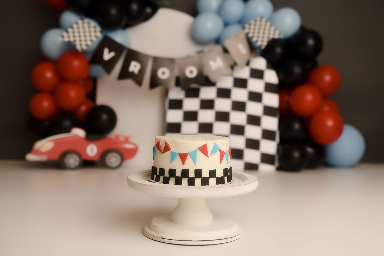 Race car themed smash cake with checkered pattern, red toy car, balloon garland, and VROOM banner for first birthday.