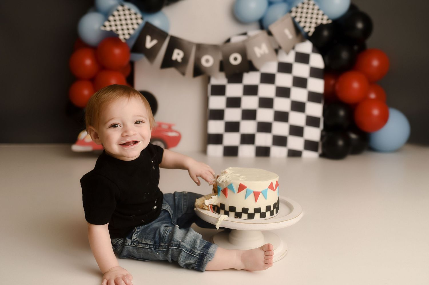 Baby boy smiling at his race car themed first birthday cake smash with checkered flag backdrop.