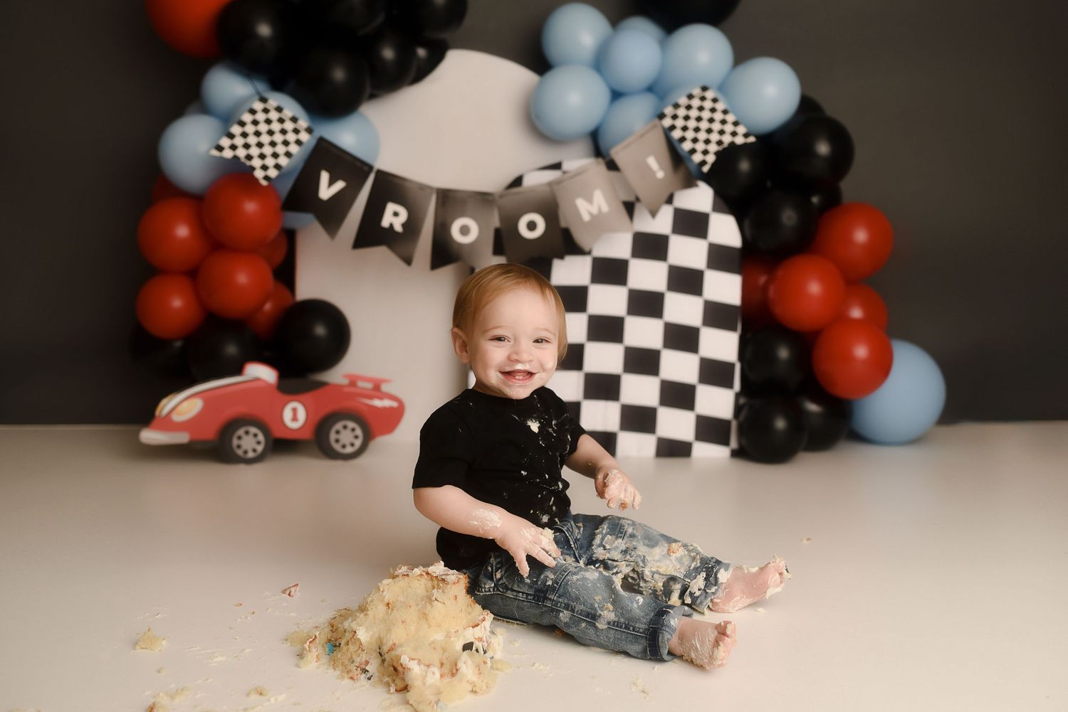 Baby smashing cake at race car themed first birthday photo session with checkered balloons and Vroom banner.